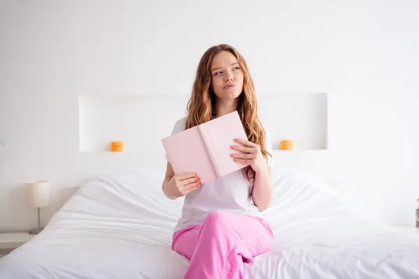 Photo of pensive minded female person sit bed hold opened book look curious away contemplate pastime modern bedroom house inside.