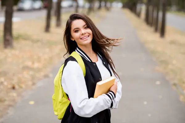 穿着制服背包抱着书的聪明漂亮的黑发女孩的画像在绿城街上的户外讲演 — 图库照片