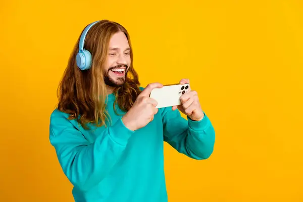 Portrait of excited cheerful man listen music headphones play games smart phone empty space isolated on yellow color background.