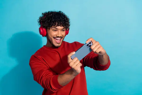 stock image Photo of youngster funny guy wear red sweatshirt playing mobile games in smartphone with headphones isolated on blue color background.