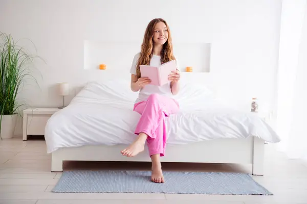 Full body portrait of positive minded girl sit comfy bed hold book look window imagine bright interior bedroom flat indoors.