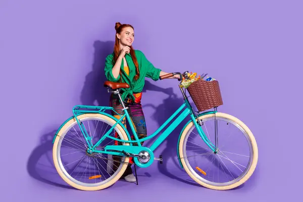 Full length photo of cheerful girl wear shirt print trousers hold bicycle look at logo empty space isolated on violet color background.