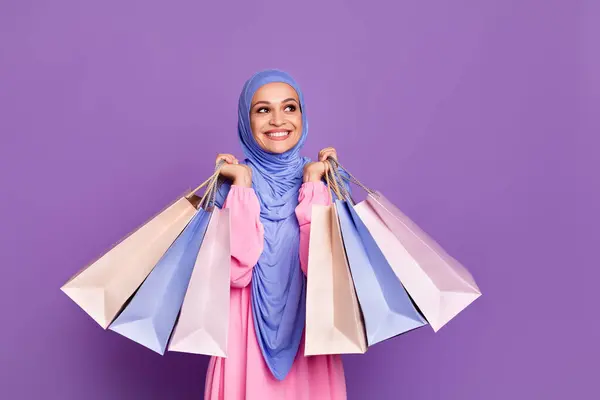 印象的な若い女性ホールドバッグの写真は 青いヘッドスカーフピンクアラビアのドレスを着用して空のスペースに見えます バイオレット色の背景に隔離 — ストック写真