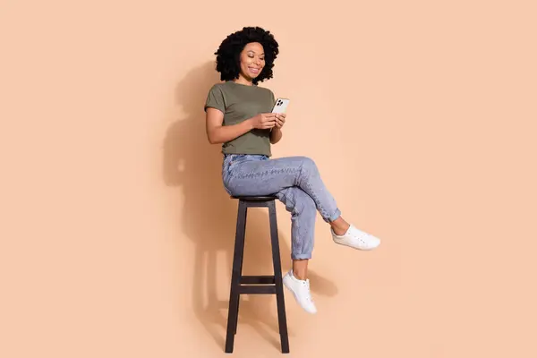 素敵な女性の完全な長さの写真は カキTシャツジーンズを着用します スマートフォンを見て ベージュ色の背景に隔離されたバースツールテキストに座って — ストック写真