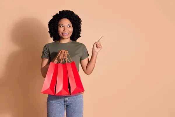 ポジティブな女性の写真は カーキTシャツを着用し ベージュ色の背景に隔離されたプロモーション空きスペースを示すショッピングバッグを見ます — ストック写真
