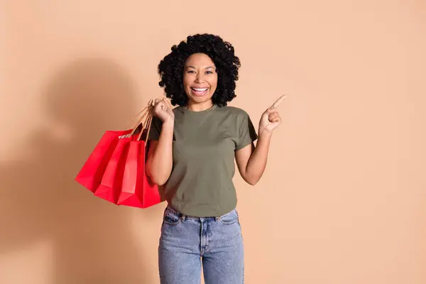 楽観的な女性の写真は カーキTシャツを着用し ベージュ色の背景に隔離された空きスペースを示すショッピングバッグを保持します — ストック写真