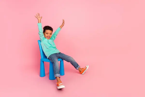 全长照片 可爱的小男孩坐在椅子上 高高兴兴地举手 穿着时髦的淡蓝色衣服 背景是粉红色的 — 图库照片