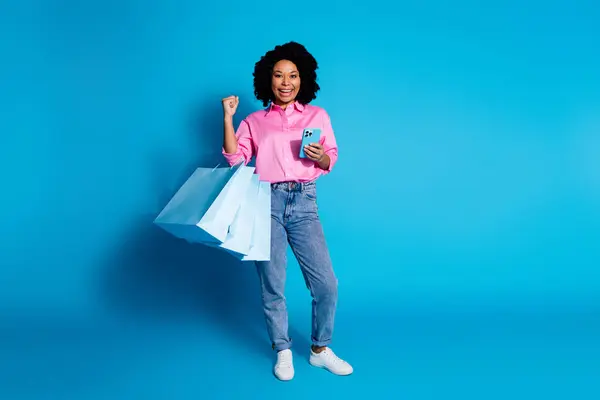 かなり若い女性のフルサイズの写真 ショップバッグを保持 携帯電話を上げる 空白スペース 青い色の背景に隔離されたピンクのシャツを着用 — ストック写真