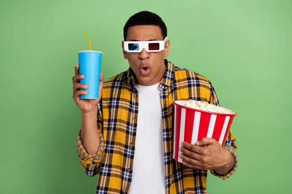 Portrait of impressed young man watch movie 3d glasses hold popcorn soda drink isolated on green color background.