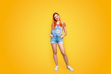 Full-body portrait of charming young girl stands isolated on bright blue background with hands in pockets of blue overalls. clipart