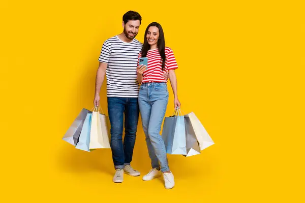 ショッピングバッグを持つポジティブなカップルの男の完全な長さの写真は 黄色の背景で隔離されたスマートフォンの読み取り通知を見ます — ストック写真