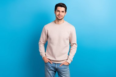 Photo of cheerful good mood man with stylish haircut dressed beige pullover hold arms in pockets isolated on blue color background. clipart