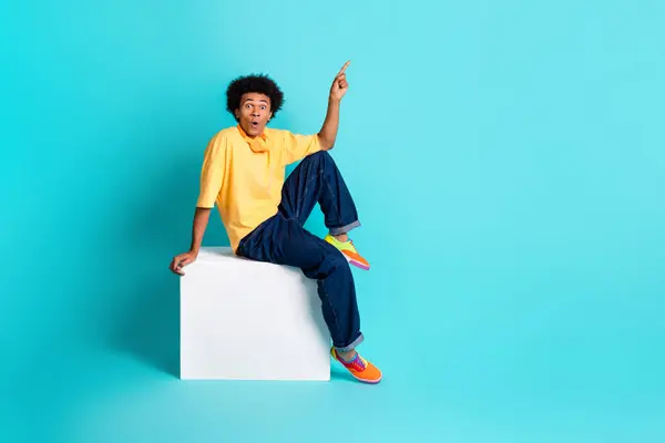 Full size photo of impressed man wear oversize t-shirt sit on cube indicating at sale empty space isolated on teal color background.