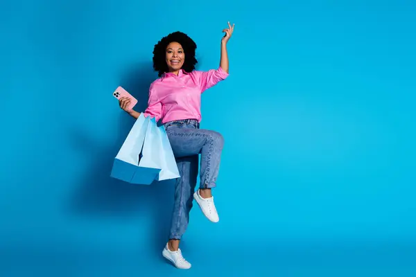 漂亮的年轻女子拿着购物袋的全长照片智能电话空旷的空间穿着蓝色背景的粉色衬衫 — 图库照片