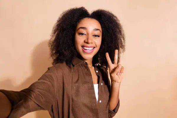 stock image Portrait of pleasant cheerful woman wavy hair dressed brown shirt showing v-sign symbol make selfie isolated on beige color background.