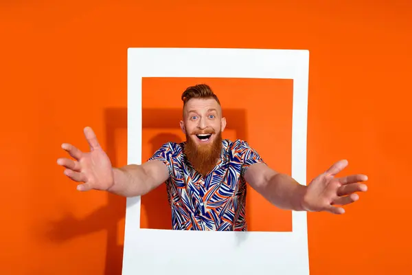Photo of eccentric guy with red beard wear print shirt in frame stretching hands catching object isolated on orange color background.