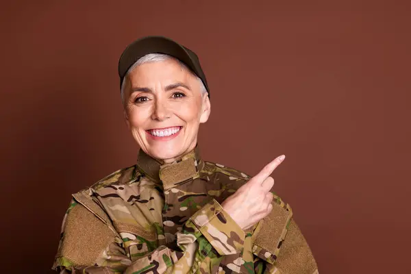 stock image Photo of lovely pensioner lady professional officer point empty space dressed camouflage army uniform isolated on brown color background.