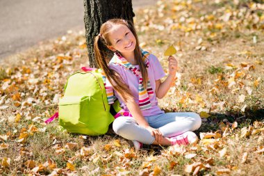 Photo portrait of ponytails pretty small girl rucksack sit under tree hold leaf wear trendy outfit elementary school student walk park. clipart