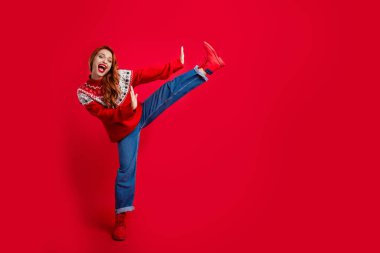 Photo of funky young redhair lady wearing ugly sweater dancing or stretching like karate fighter isolated on red color background. clipart