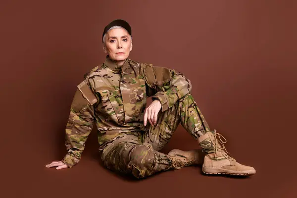 stock image Full body photo of attractive mature woman army soldier sit floor relaxed wear military uniform isolated on brown color background.