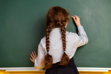 Rear portrait of little schoolchild girl write chalk blackboard wear uniform isolated on yellow color background. clipart