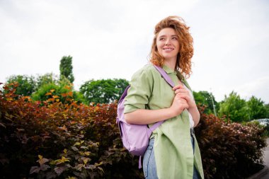 A cheerful young red-haired woman with a backpack enjoys a sunny day outdoors, surrounded by greenery, embodying joy and relaxation. clipart