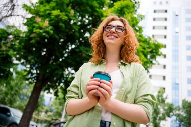 A young redhaired woman with glasses enjoys a sunny day outdoors, holding a coffee cup, showing a relaxed smile, while promenading in a green city park. clipart