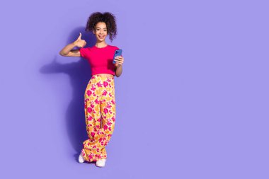 Full length photo of charming positive woman wear pink top showing thumb up chatting modern gadget empty space isolated violet color background. clipart