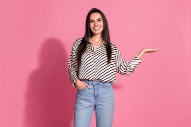 A young brunette woman in a striped shirt and jeans smiles confidently while gesturing against a pink background, embodying casual style and positive vibes. Perfect for advertising and promoting. clipart