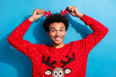 Photo portrait of handsome young guy show touch deer headband dressed red christmas sweater celebrate winter holidays on blue background. clipart