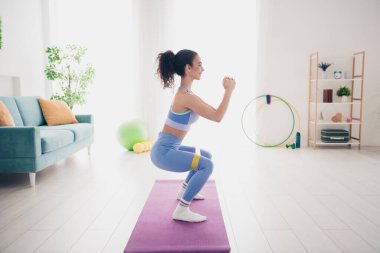 A young woman in trendy activewear performs an indoor workout on a yoga mat, surrounded by a bright, modern living room setup with natural light and plants. clipart