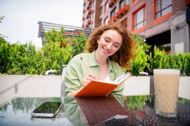 Young redhaired woman sits outdoors in natural light, joyfully writing in a journal, sipping coffee, and enjoying a sunny day. clipart