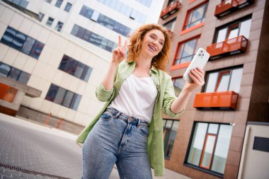 A young redhaired woman enjoying a sunny day outdoors, taking selfies with her smartphone in the city, embodying joy and leisure. clipart