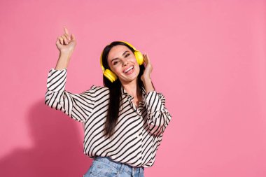 Joyful young brunette woman wearing striped shirt with yellow headphones, dancing against a vibrant pink background, showcasing happiness and enjoyment. clipart