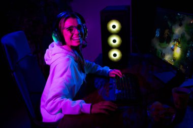 A cheerful young female gamer in casual wear enjoying a video game session at night, illuminated by neon lights, creating a relaxed and happy atmosphere at home. clipart