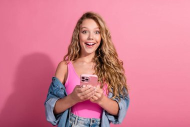 Joyful young female in fashionable denim holding smartphone against a vivid pink background, radiating happiness and charm. clipart