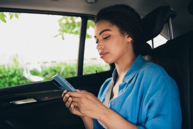 A young woman relaxes in a car, using her smartphone during a sunny day in the city. Her casual style and happy expression reflect a carefree moment. clipart