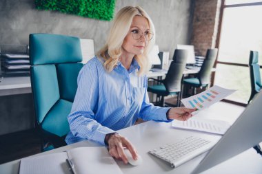 Mature professional woman working in a bright office, examining graphs and data on a computer. She wears elegant attire and exudes confidence, representing successful business dynamics and modern clipart