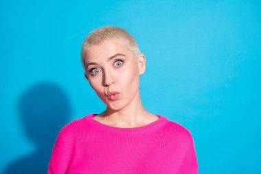 A young woman with short blond hair makes a surprised face against a vivid blue background, wearing a pink sweater that matches the fashionable vibe. clipart