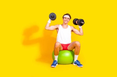 Cheerful young man in sportswear lifting dumbbells, sitting on gym ball against vivid yellow backdrop, epitomizing a healthy and active lifestyle. clipart