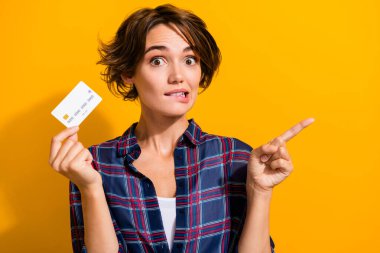 Photo of attractive woman dressed shirt hold credit card indicating at promo empty space biting lips isolated on yellow color background. clipart