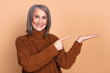 Cheerful mature woman in a brown sweater smiling and pointing to her extended hand, set against a simple beige background, invoking warmth and approachability. clipart