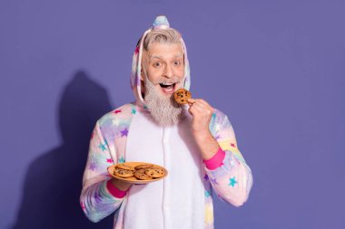 A cheerful mature man in a stylish unicorn onesie enjoys cookies, radiating joy and positivity against a purple background, perfect for advertising or lifestyle promotion. clipart