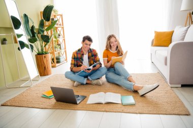 A happy young couple sitting on the floor, studying together. Surrounded by books and a laptop, they enjoy a relaxed weekend at home in a bright, cozy living room. clipart