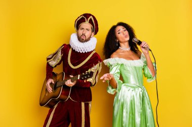 Two individuals dressed as a royal couple in creative costumes, singing and playing music against a vibrant yellow background. Features a classical touch with a vivid modern setting. clipart