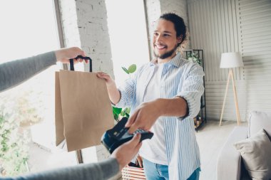 A cheerful man in casual wear receiving a delivery paper bag at home in a bright, cozy living room setting clipart