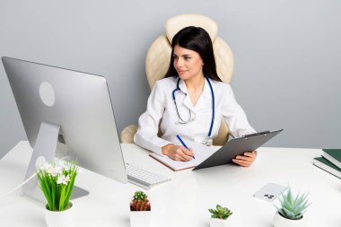 A female doctor in a white coat with a stethoscope reviewing documents while seated at a desk in a clinic office, showcasing professionalism and healthcare ambiance. clipart