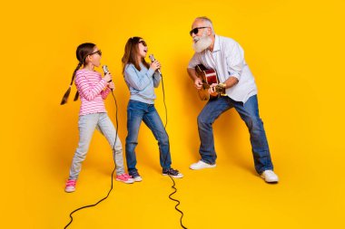 A cheerful elderly man plays guitar alongside two joyful young girls singing together, set against a vivid yellow background, embodying family connection, happiness, and a lively atmosphere of shared clipart