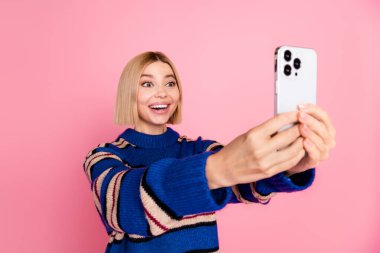 A cheerful young woman with a stylish bob haircut holding a smartphone for a selfie. She is dressed in a fashionable striped sweater and stands against a vibrant pink background, exuding charm and clipart