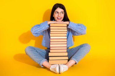 Smiling young woman leaning on a tall pile of books on a bright yellow background, wearing casual stylish attire, showcasing a cheerful and carefree lifestyle. clipart